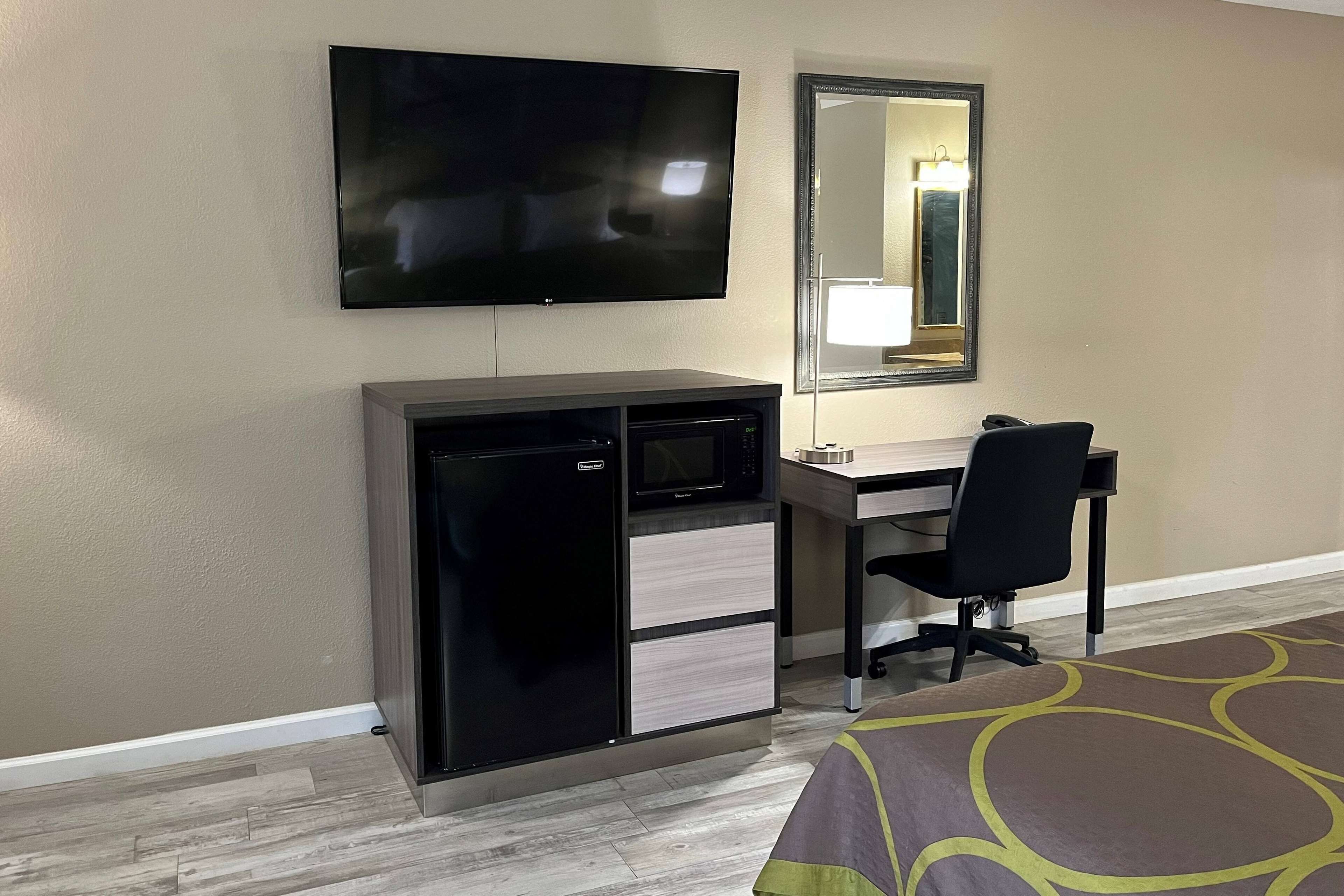 A hotel room with a flat-screen TV mounted on the wall, a compact refrigerator, a desk with a chair, and a large mirror.
