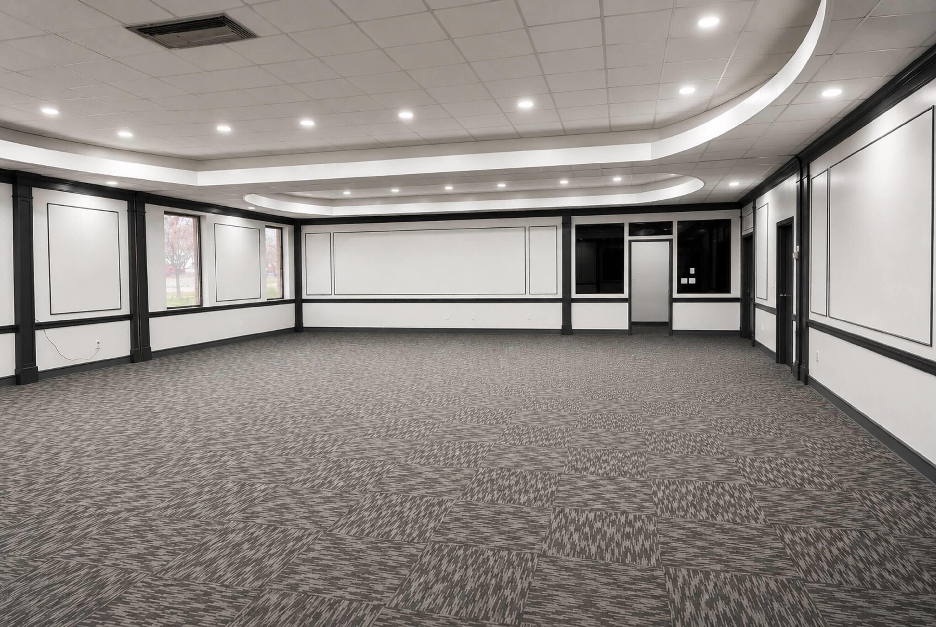 Un'ampia sala conferenze vuota, con tappeto a motivi, pareti bianche con modanature nere e illuminazione a soffitto incassata.