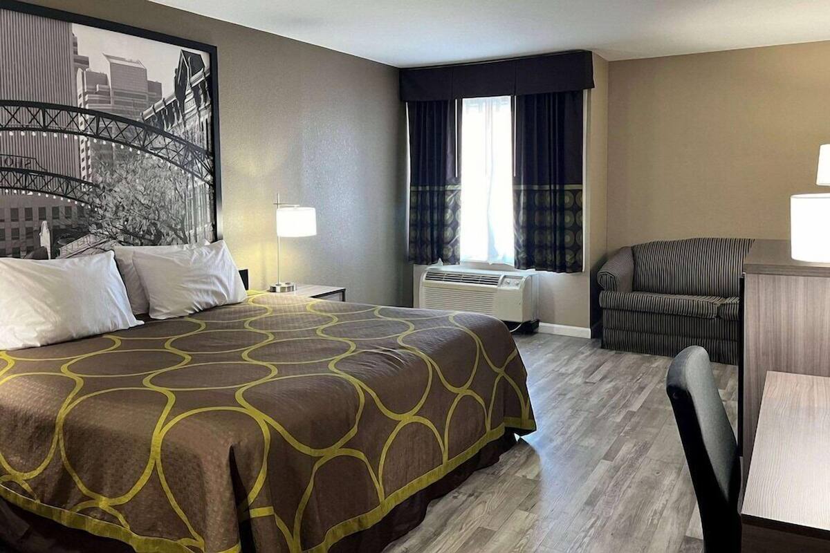 Une chambre d’hôtel avec un lit, un bureau, une chaise, une fenêtre avec des rideaux, un climatiseur et un tableau représentant un paysage urbain accroché au mur.