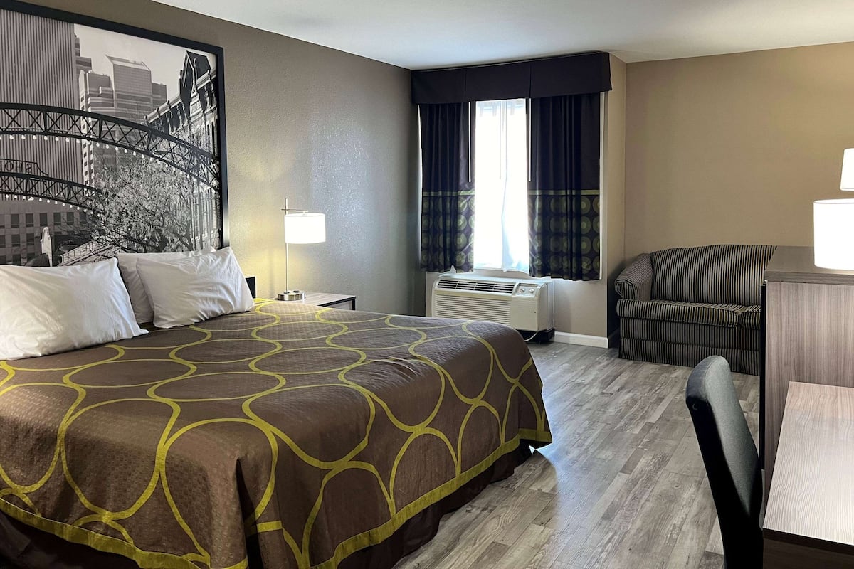 Une chambre d’hôtel avec un grand lit, un bureau avec une chaise, un appareil de climatisation et un tableau représentant un paysage urbain accroché au mur.