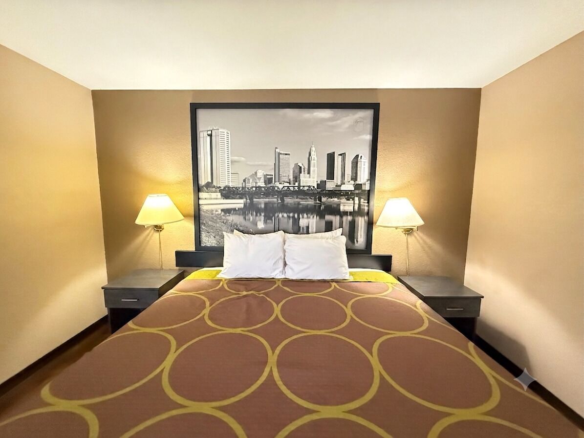 A hotel room with a bed, two bedside tables, a cityscape painting on the wall, and two lamps.