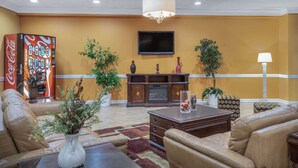 A lobby with leather sofas, a coffee table, a TV mounted on the wall, a Coca-Cola vending machine, and decorative plants. - Super 8 by Wyndham Harrison OH (Harrison)