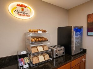 A breakfast station with a sign, a toaster, a refrigerator, and a variety of bread and pastries. - Super 8 by Wyndham Harrison OH (Harrison)