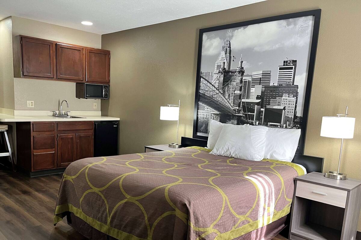 Une chambre d’hôtel comprenant un lit, une table de chevet, une lampe, un bureau avec une chaise, un four à micro-ondes et une kitchenette équipée de placards et d’un évier.