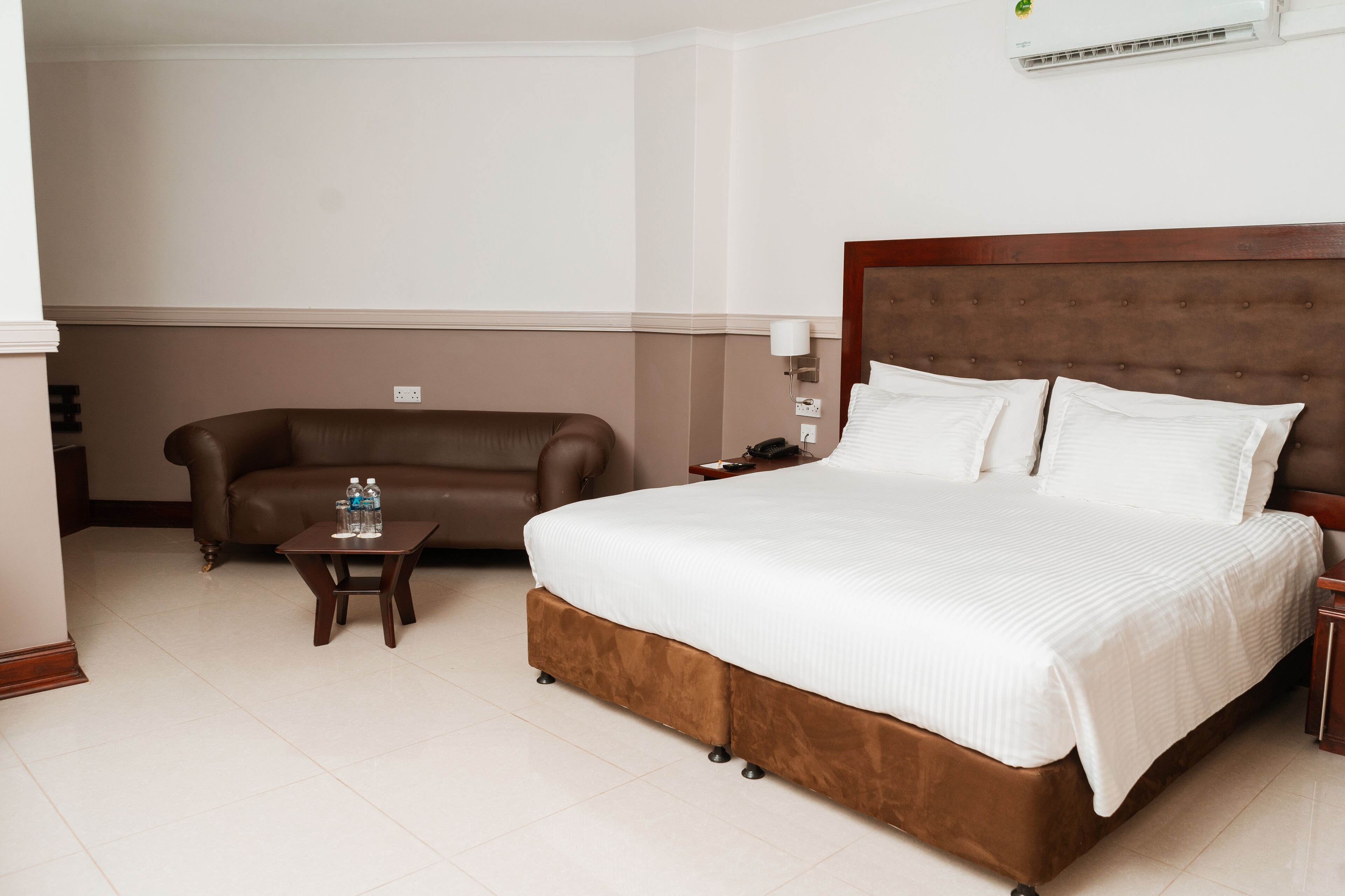 A hotel room with a large bed, a brown leather sofa, a small table with a water bottle, and a nightstand with a lamp.
