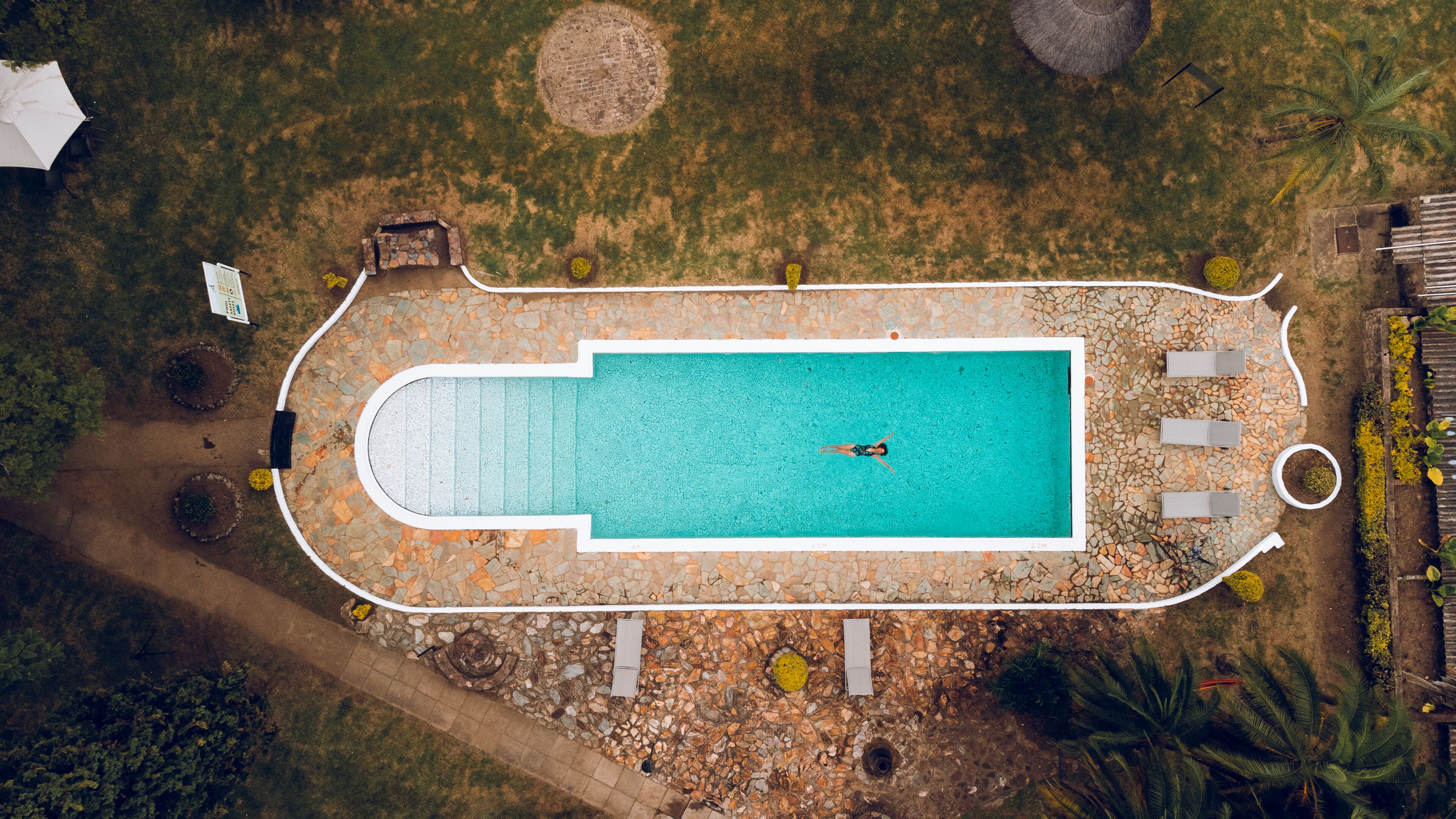An aerial view of a rectangular swimming pool with a person swimming, surrounded by a pebble-lined deck and various landscaping features.