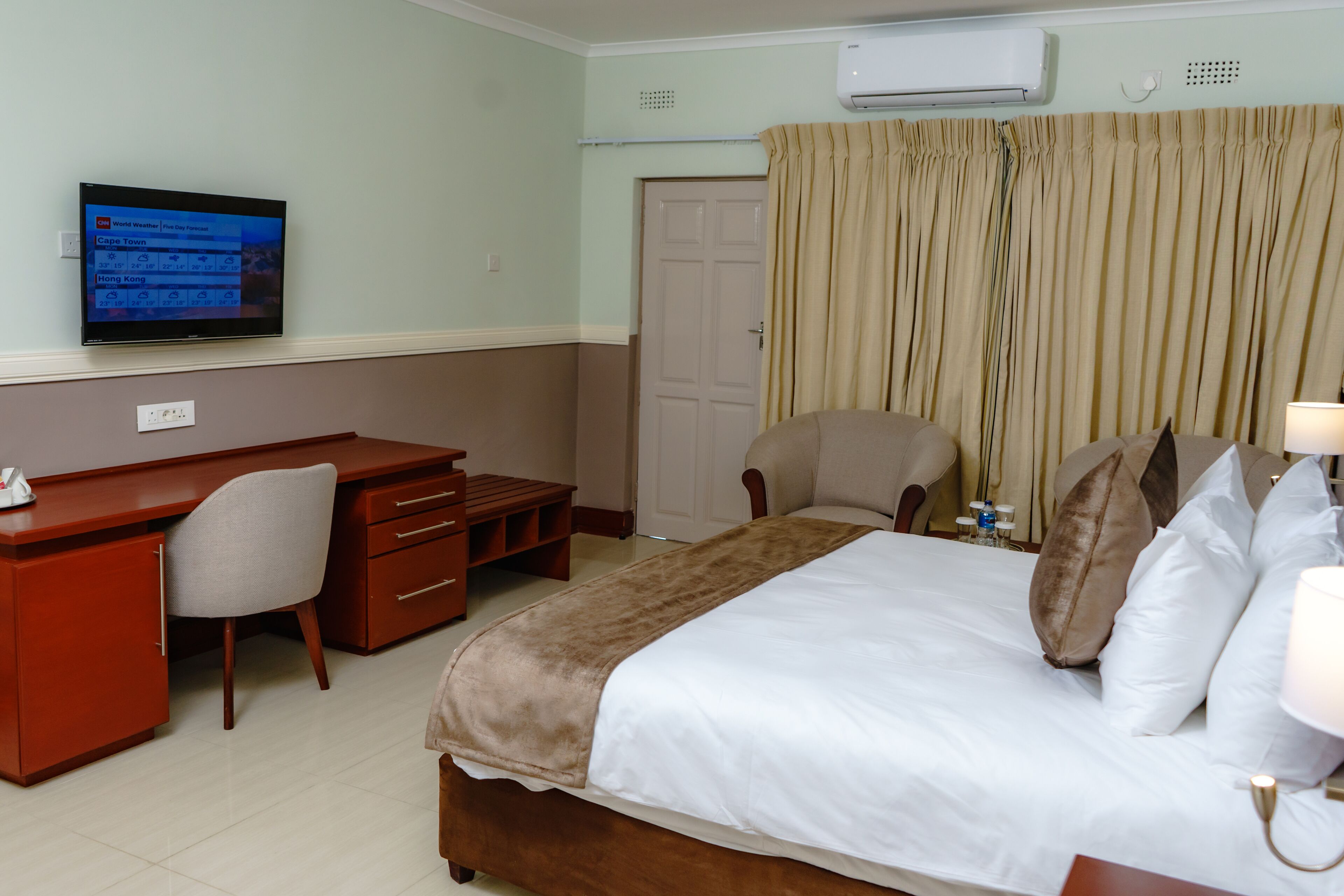 A hotel room with a bed, a desk, a chair, a TV displaying weather information, and an air conditioning unit.