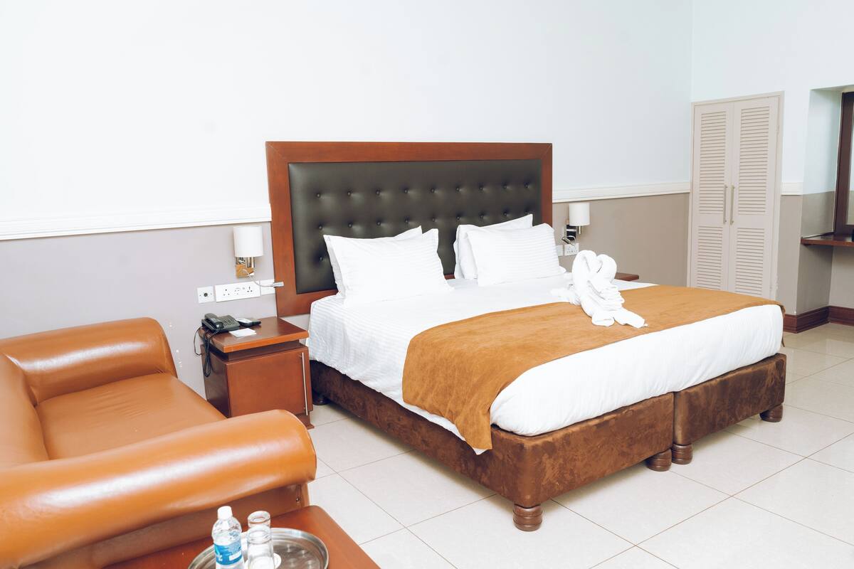 A hotel room with a large bed, a wooden headboard, a brown leather armchair, a bedside table with a lamp, and a wardrobe.