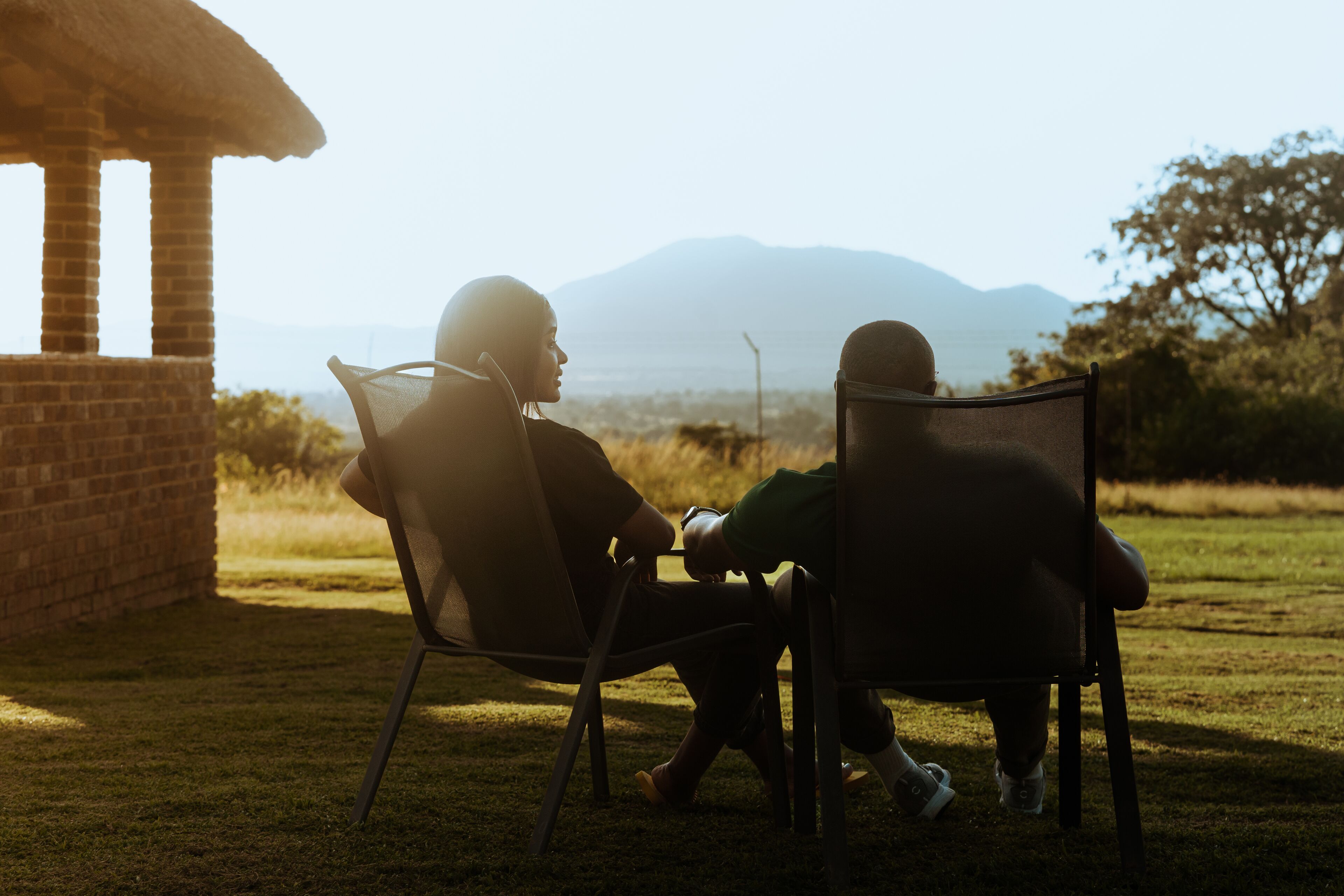 Two people sitting on chairs facing a brick structure with a view of mountains.