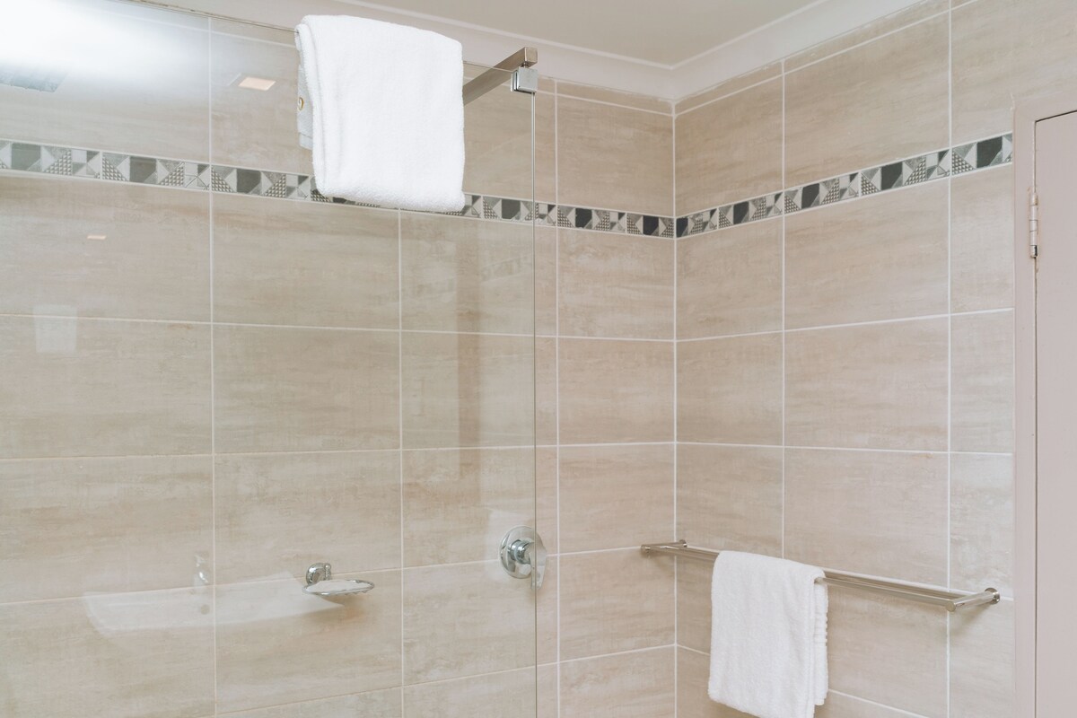 A shower with beige tiles, a glass-enclosed showerhead, and two towel racks with white towels.