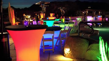 Área de piscina com mesas e cadeiras iluminadas, piscina e um cenário montanhoso.