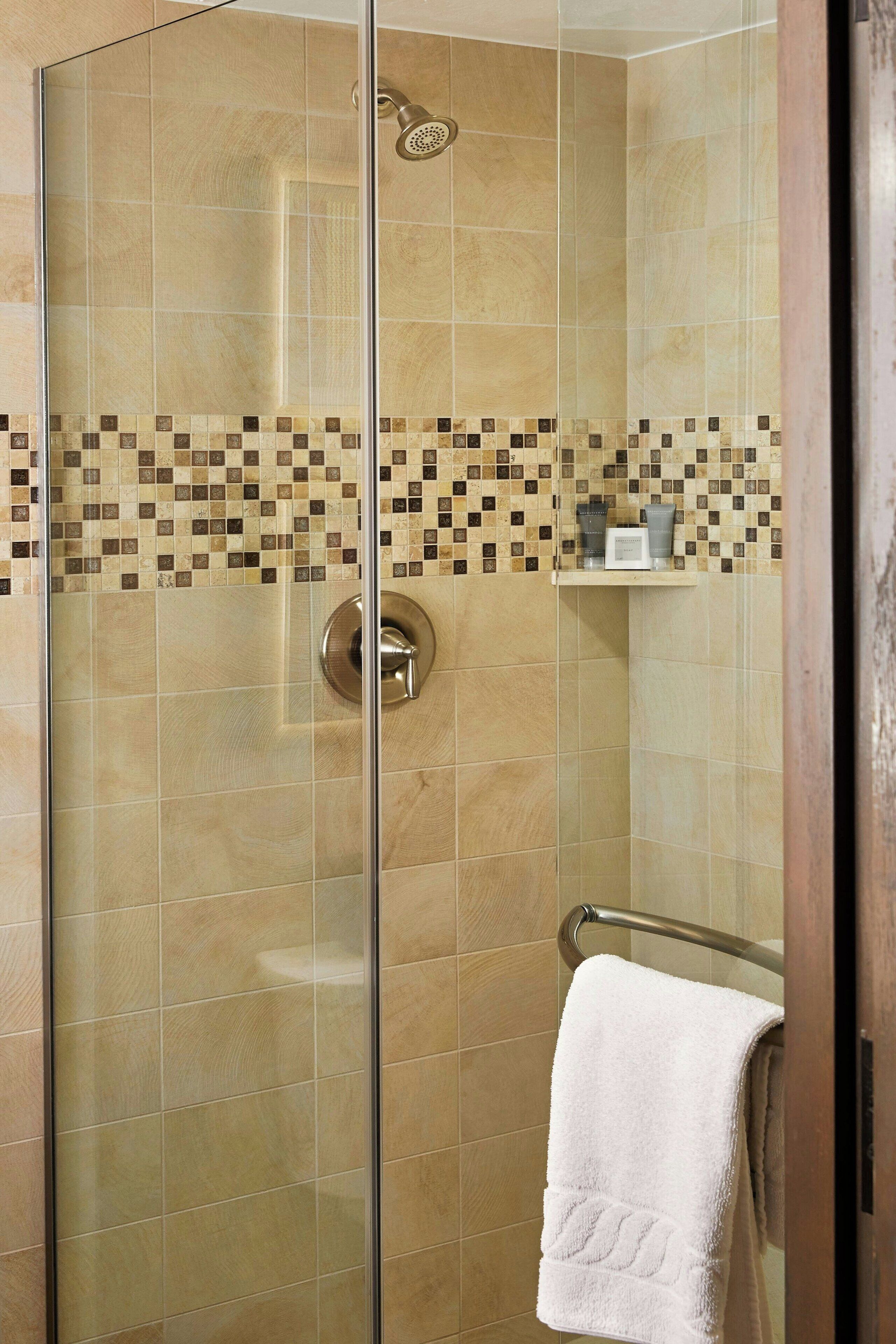A glass-enclosed shower with a tiled wall, a towel rack, and a shelf with toiletries.
