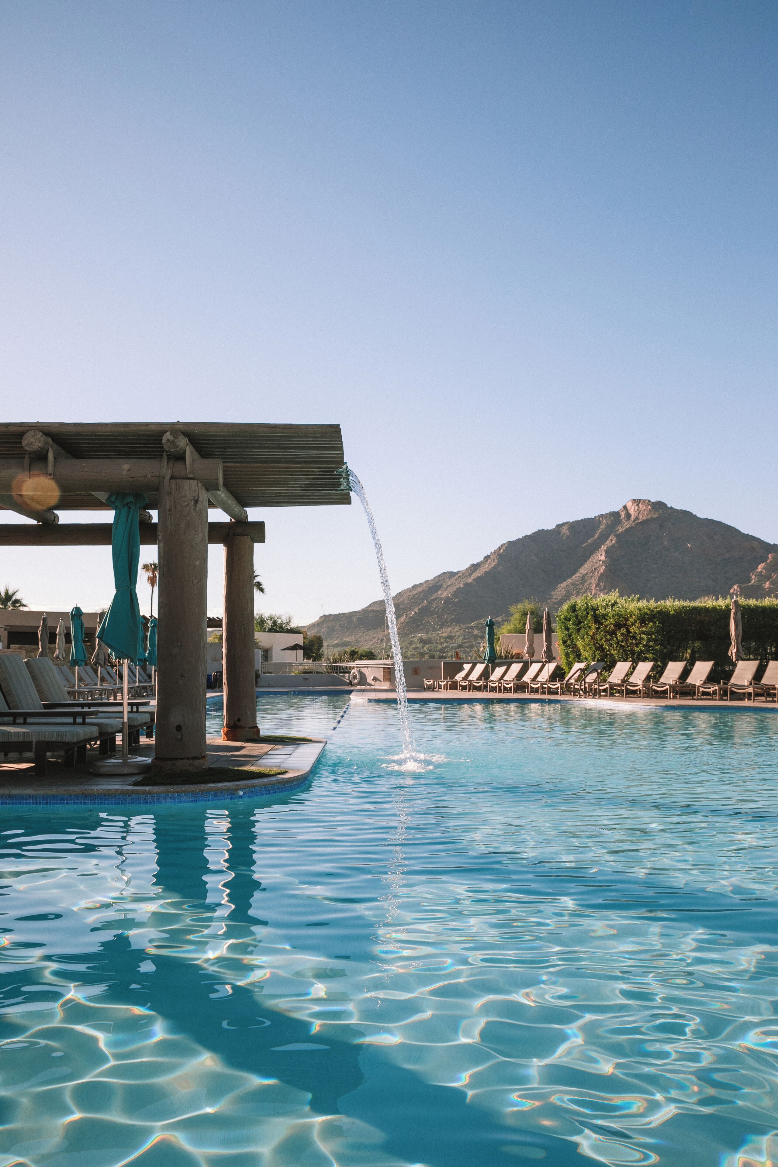 Una piscina azul cristalina con una fuente, sillas de descanso y una montaña al fondo.