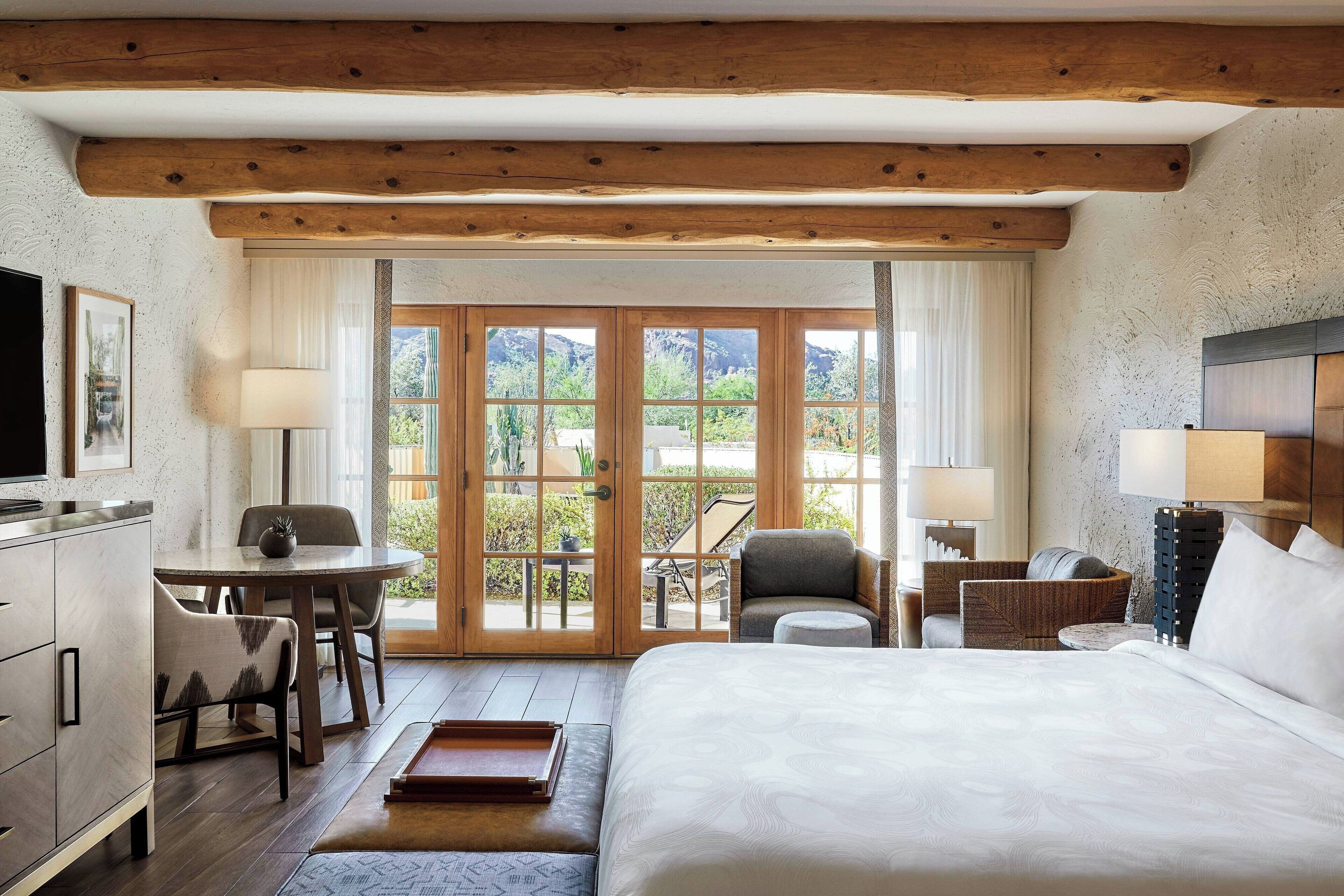 A hotel room with a large bed, a desk with a chair, a small table with a lamp, and a sofa. There are large windows with a view of greenery outside.