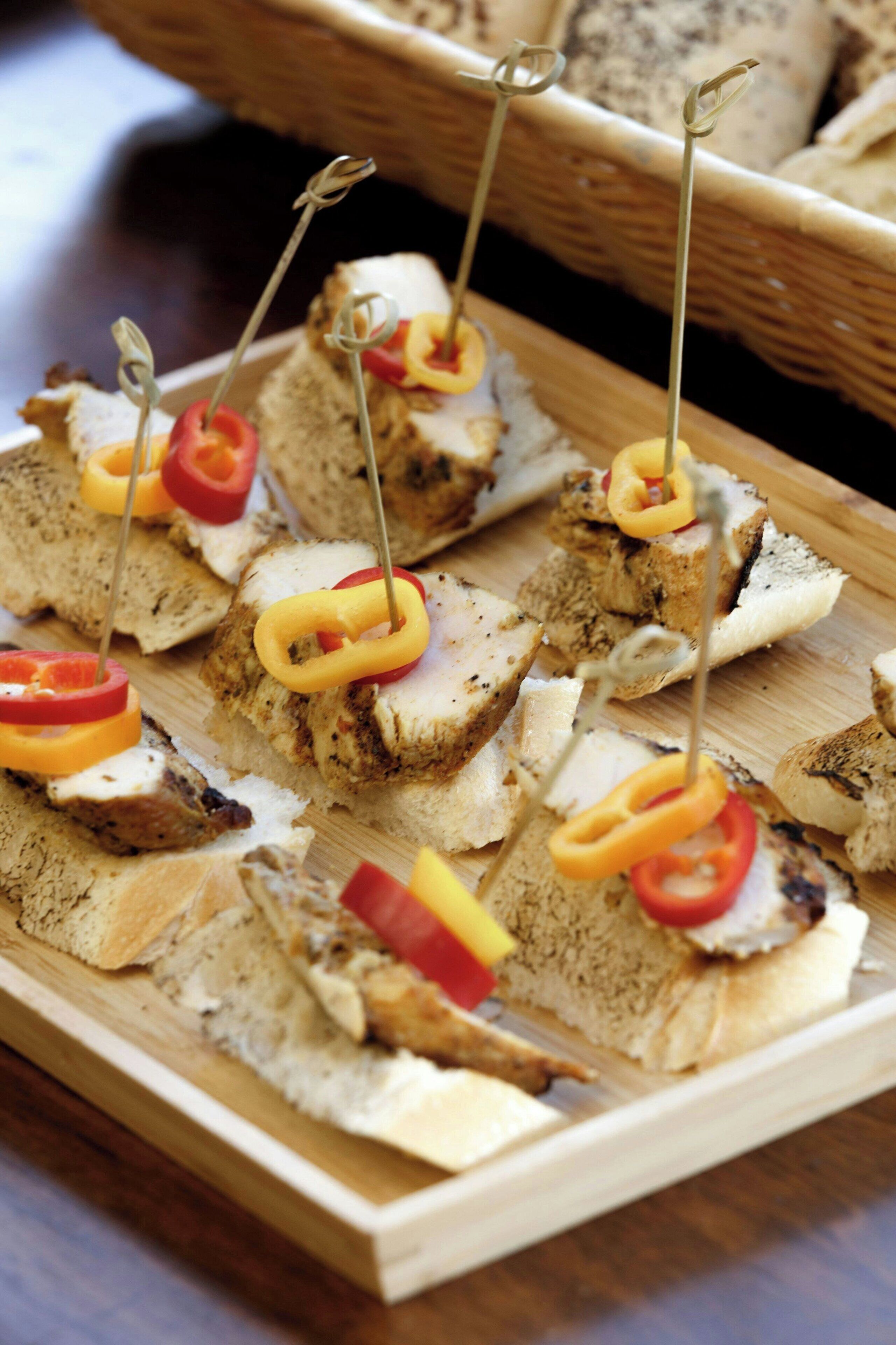 A wooden board with several pieces of toasted bread topped with slices of red and yellow bell peppers, skewered with toothpicks.