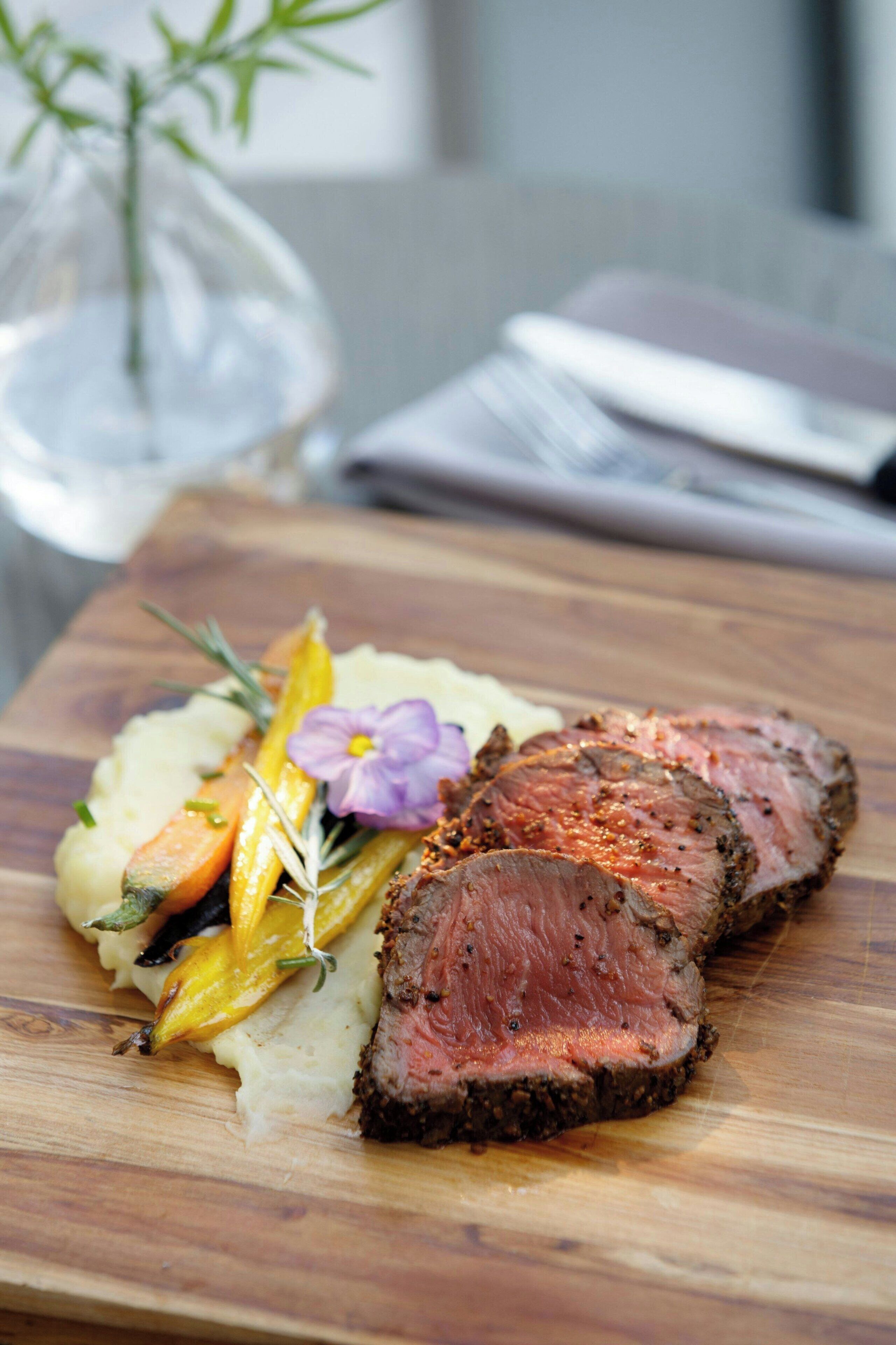 Una tabla de madera con un trozo de carne, puré de papas y verduras asadas.