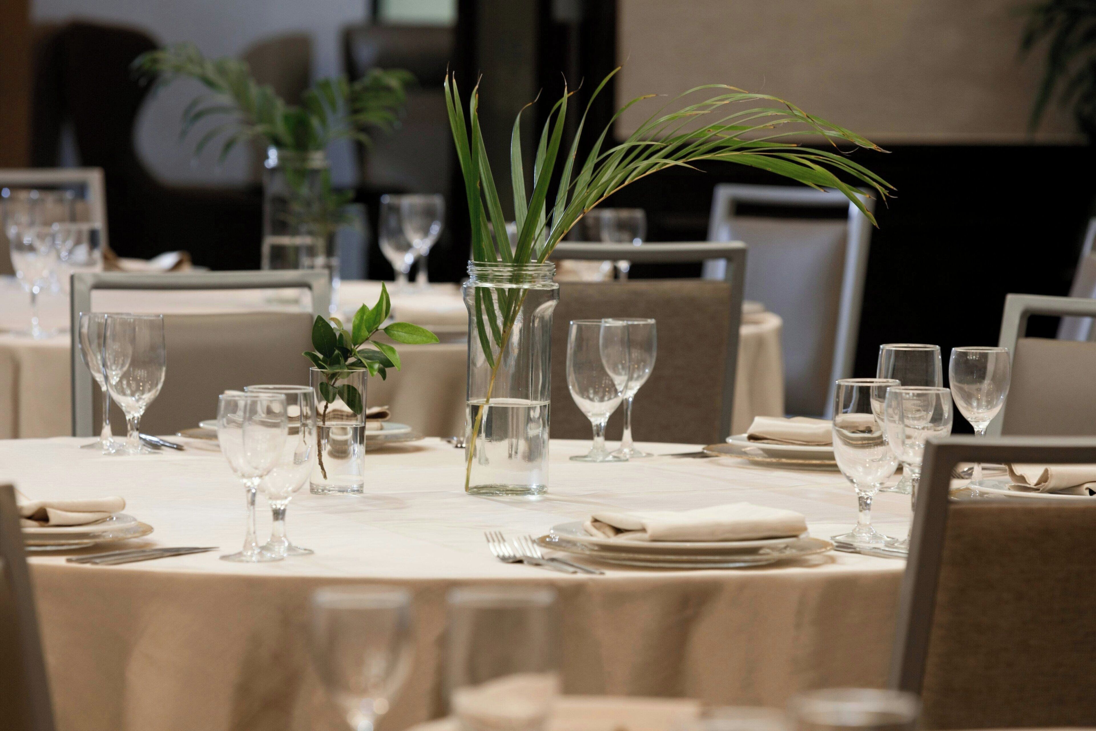 Una mesa de comedor dispuesta con copas de vino, platos y cubiertos, con un jarrón con plantas verdes en el centro.