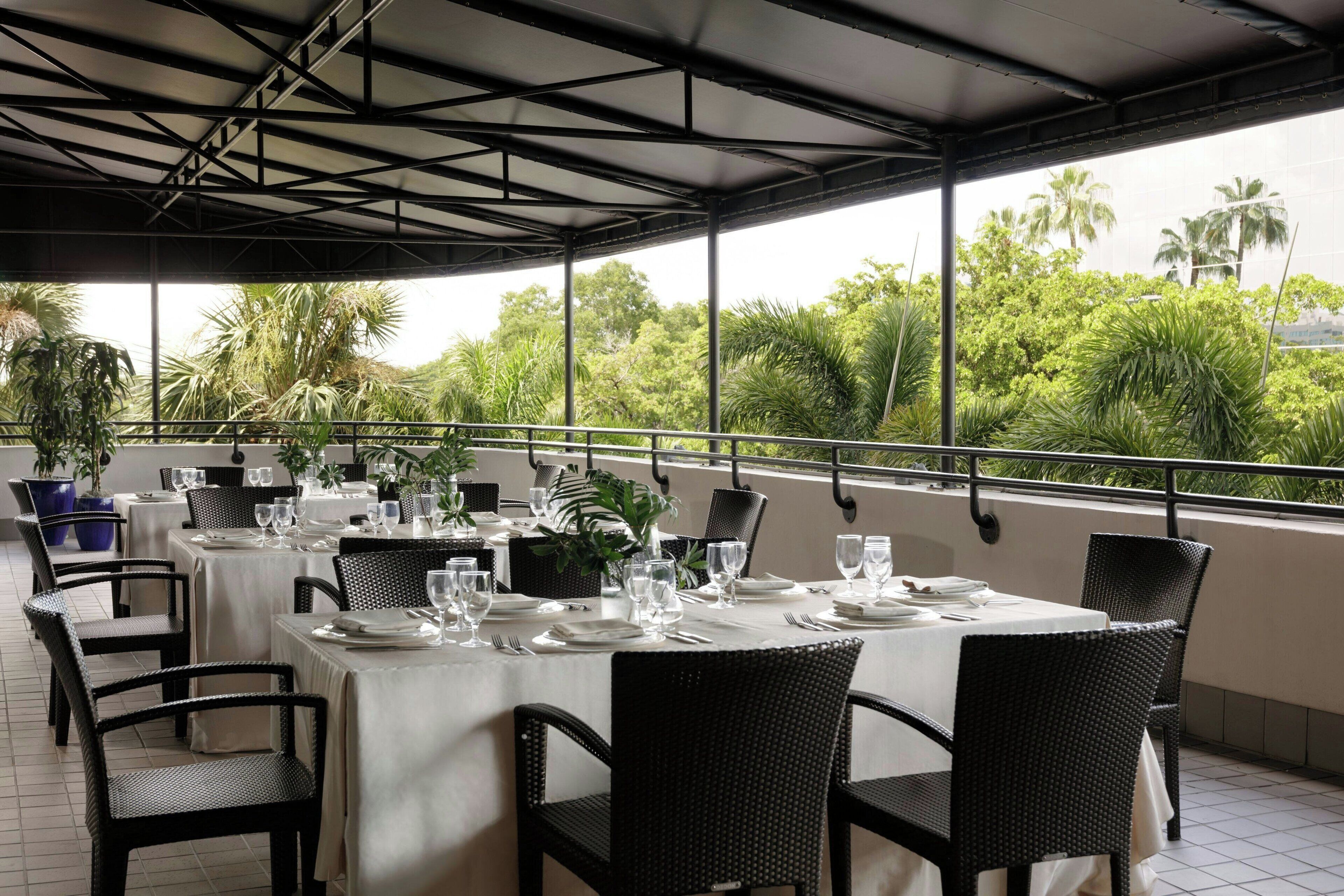 Un área de comedor al aire libre con mesas dispuestas para dos, rodeada de sillas de mimbre negro, y vistas a exuberante follaje y palmeras.
