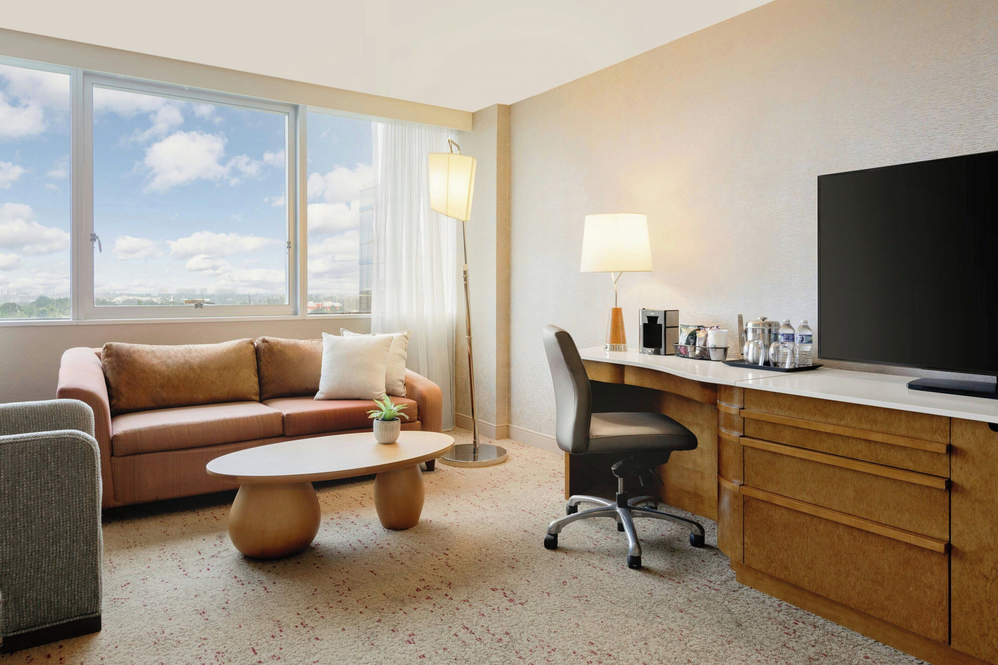 A hotel room with a sofa, a coffee table, a desk with a lamp, a chair, and a TV.