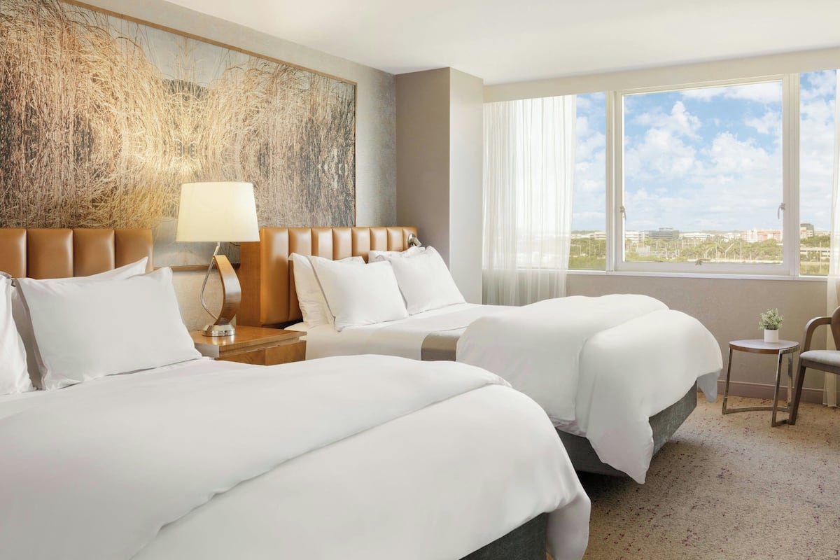 A hotel room with two beds, a large abstract painting, a window with a city view, and a small table with a lamp.