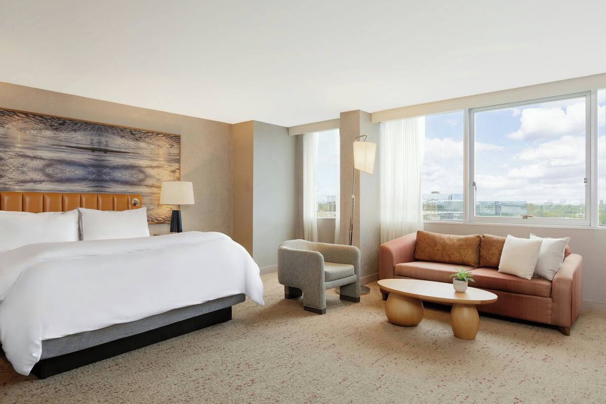 A hotel room with a large bed, a sofa, a small armchair, a coffee table, and a view of the city through large windows.