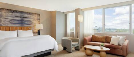 A hotel room with a large bed, a sofa, a small armchair, a coffee table, and a view of the city through large windows.