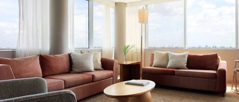 A modern hotel room with two sofas, a coffee table, and large windows offering a city view.