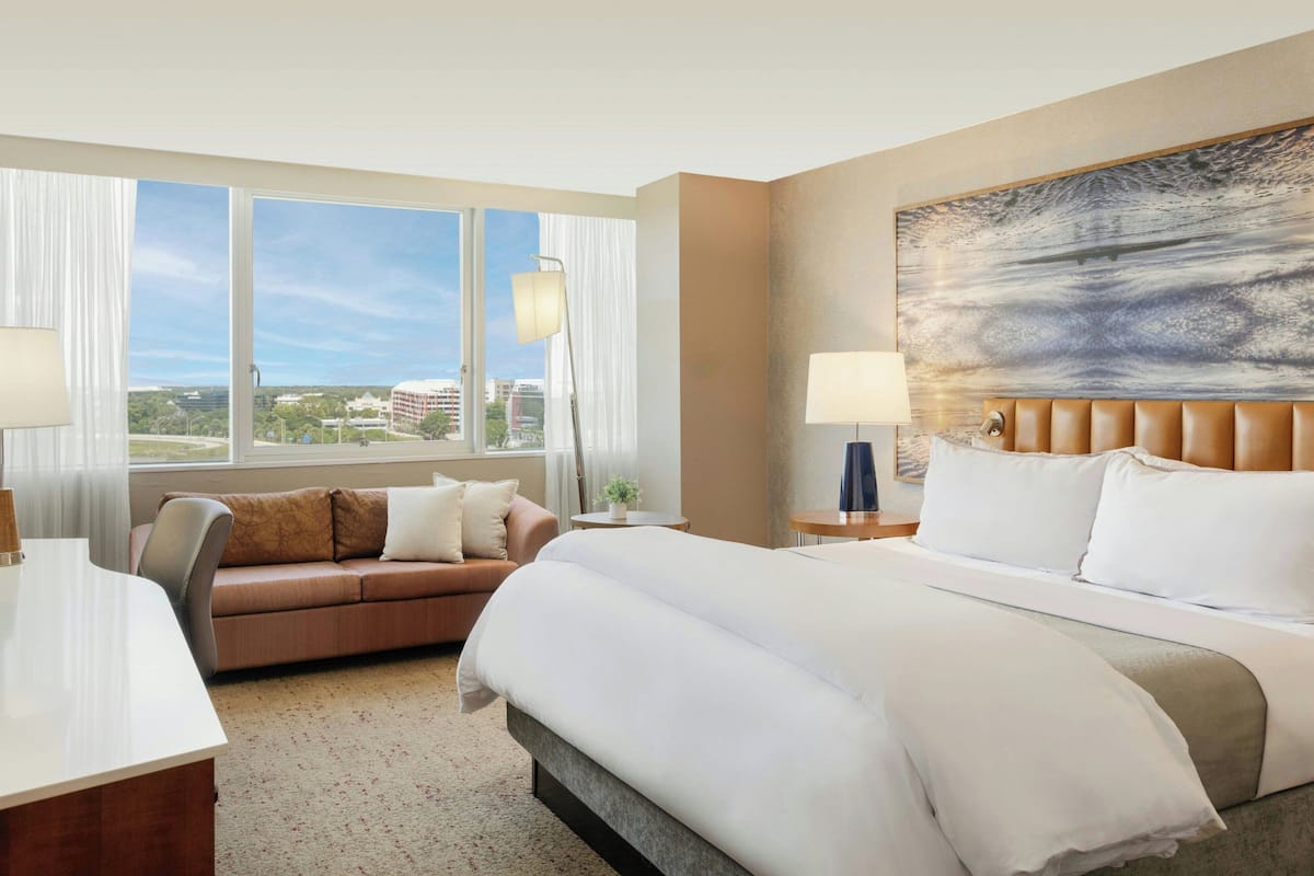 A hotel room with a large bed, a sofa, a desk, and a view of the city through large windows.