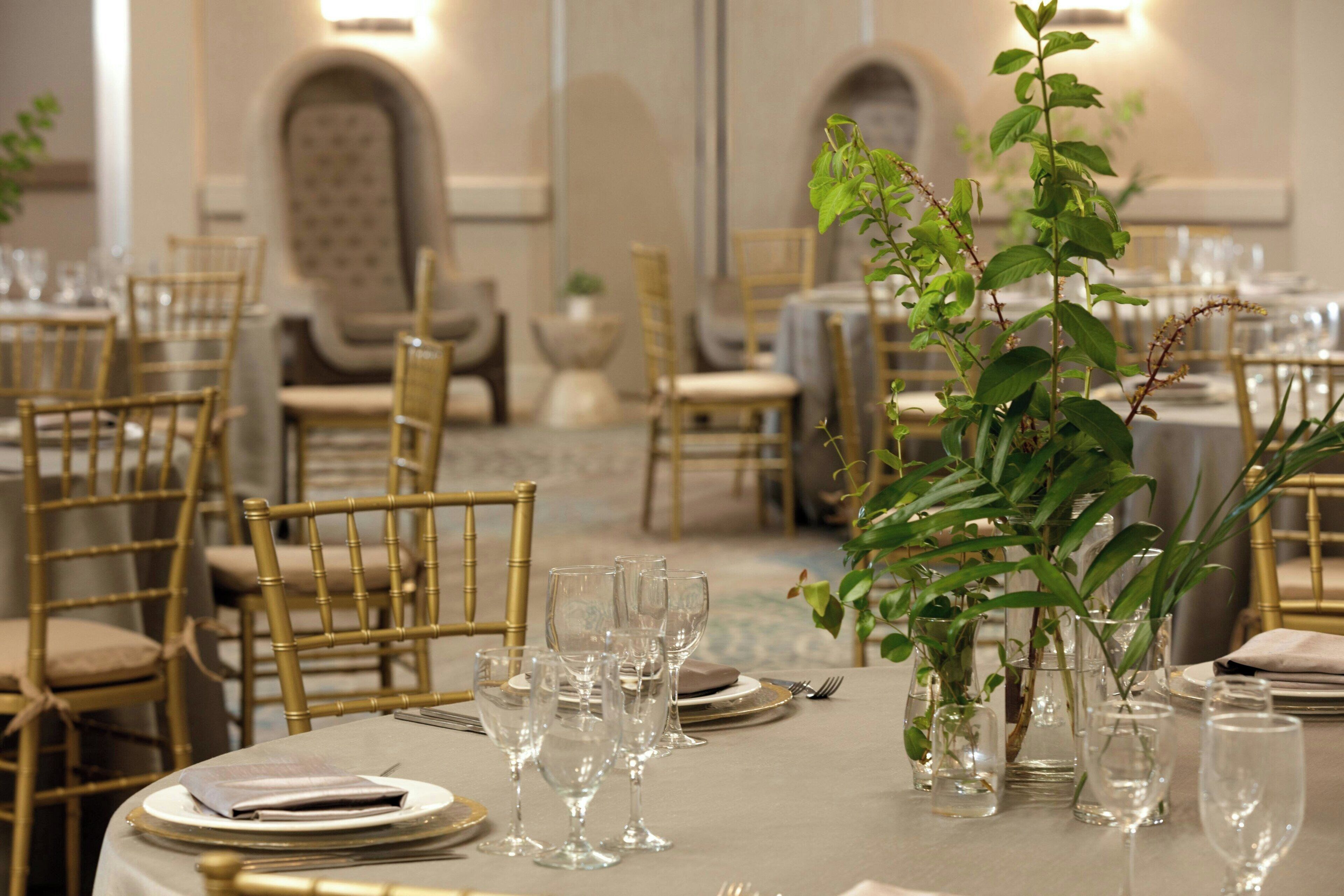 Un espace repas avec des tables rondes dressées pour le service, agrémenté de chaises dorées, de verres transparents et de petites plantes en pot.