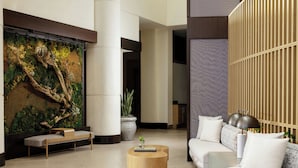 A modern lobby with a large wall-mounted green wall, a wooden bench, a round wooden coffee table, a sofa with white cushions, and a tall column.