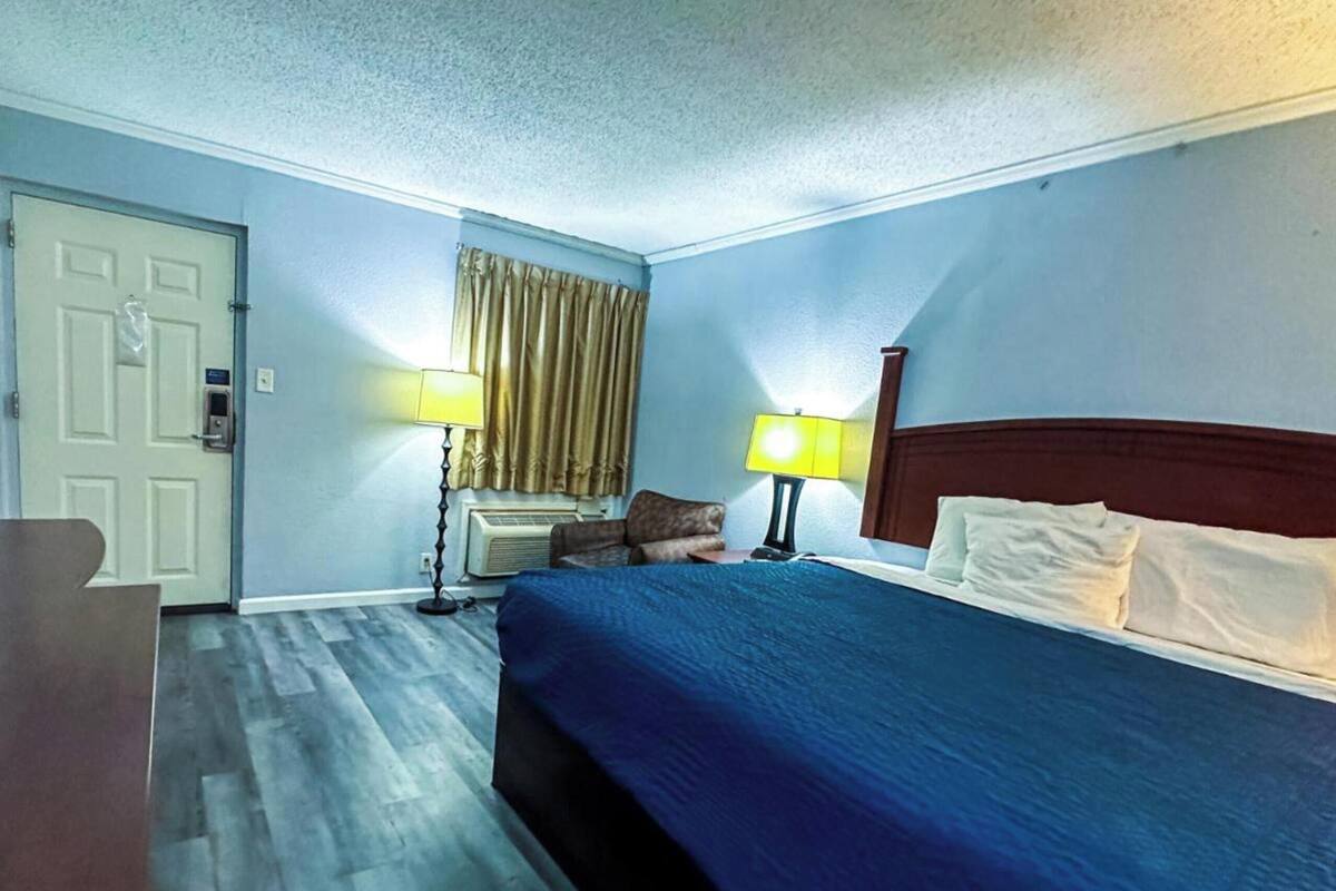 A hotel room with a bed, two bedside lamps, a window with curtains, a door, and a small table with a lamp.