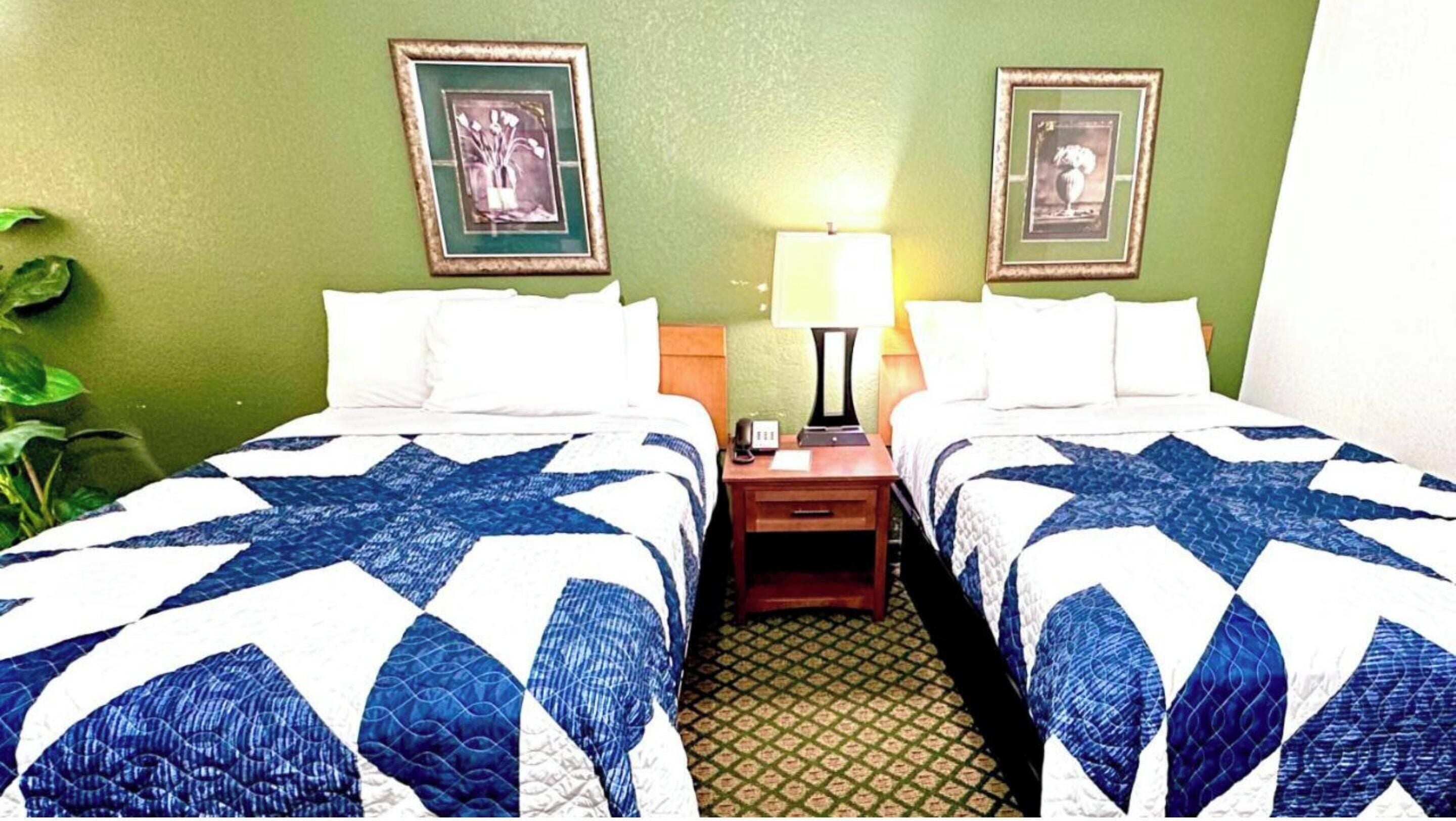 Habitación de hotel con dos camas, cada una con colcha azul y blanca, una mesita de noche con lámpara y dos cuadros enmarcados en la pared.