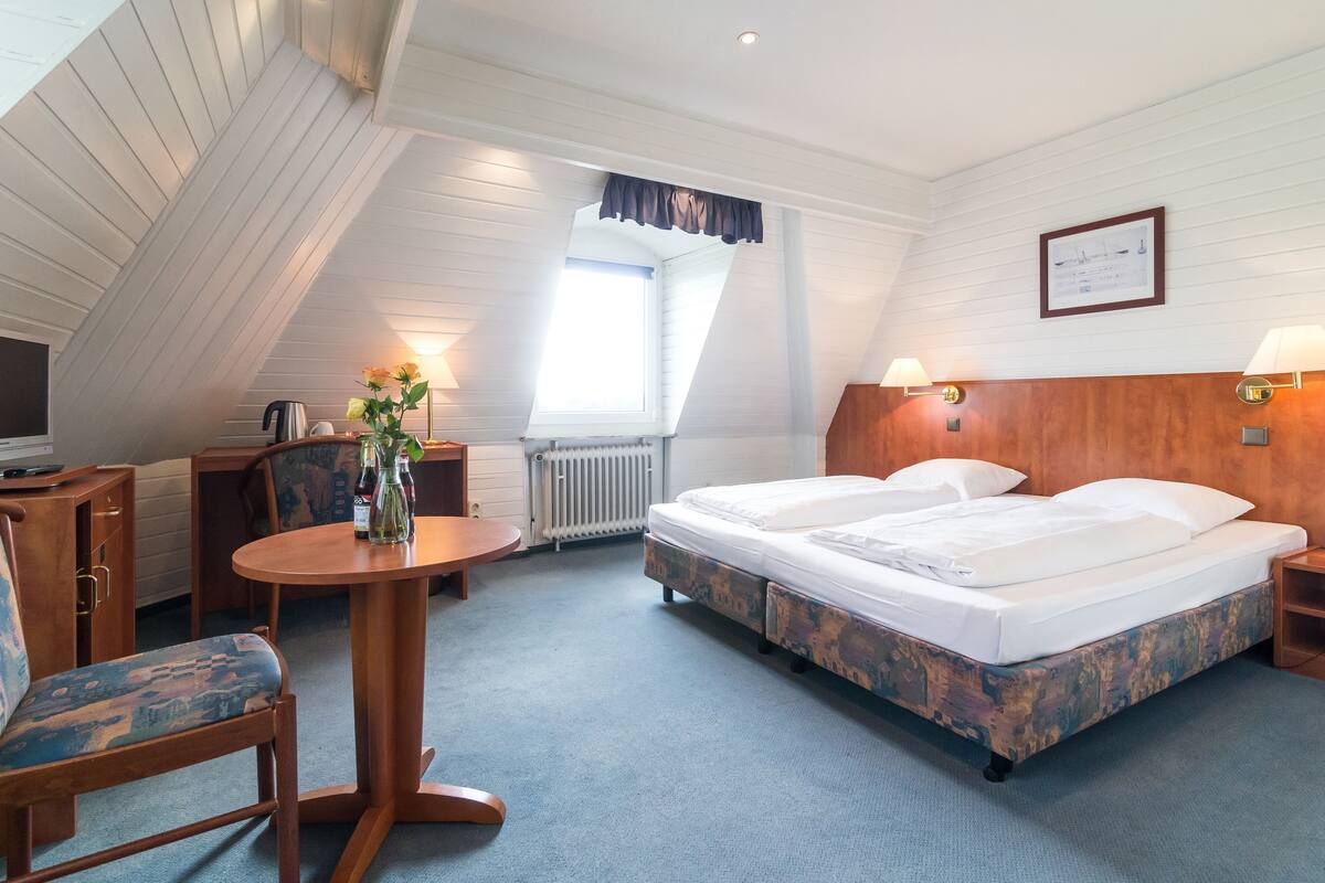 A hotel room with a loft-style ceiling, a bed with a wooden frame, a desk with a chair, a small table with a vase, and a window with curtains.