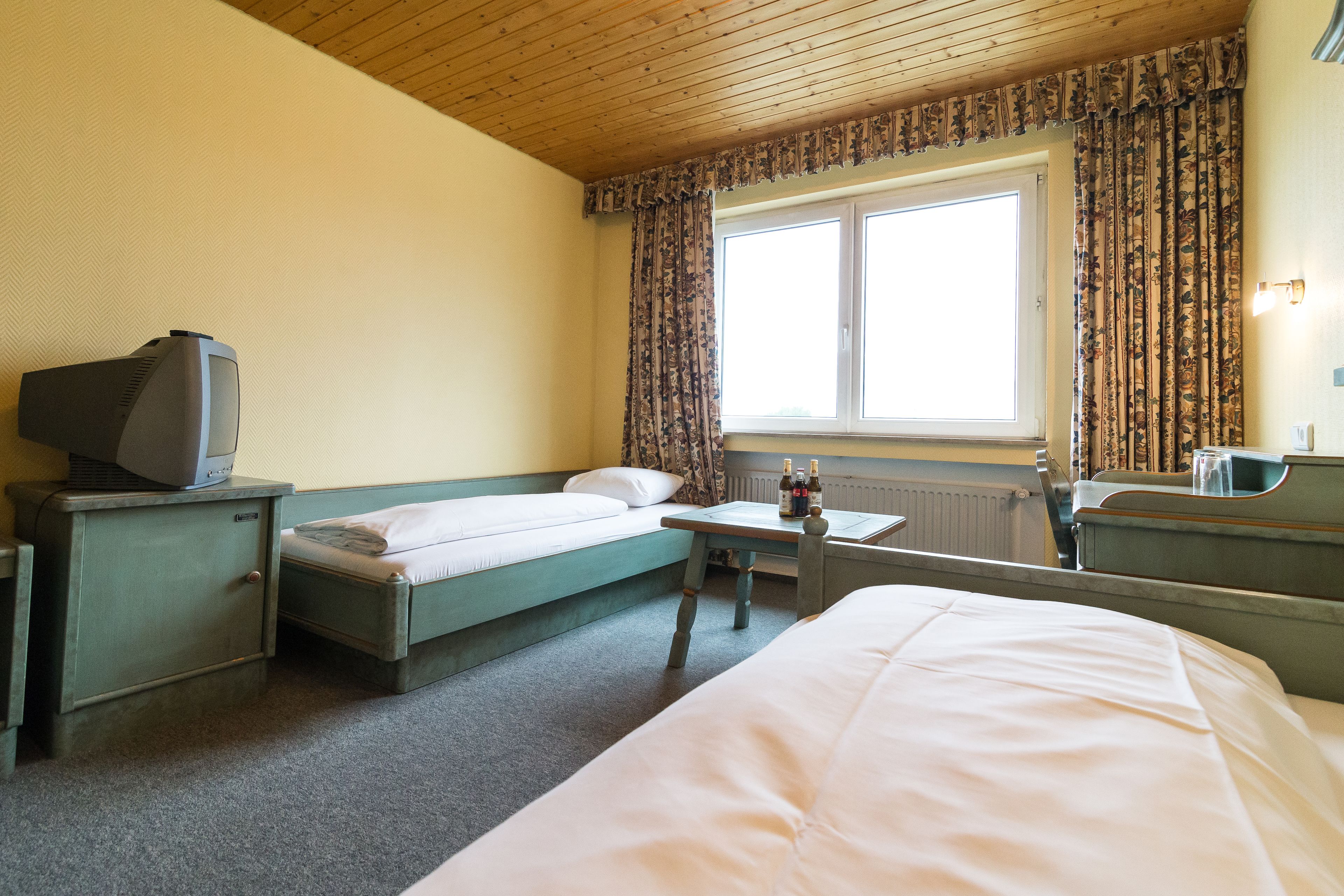 Ein schlichter, kleiner Raum mit einem Bett, einem Schreibtisch, einem Fernseher und einem Fenster mit Vorhängen.