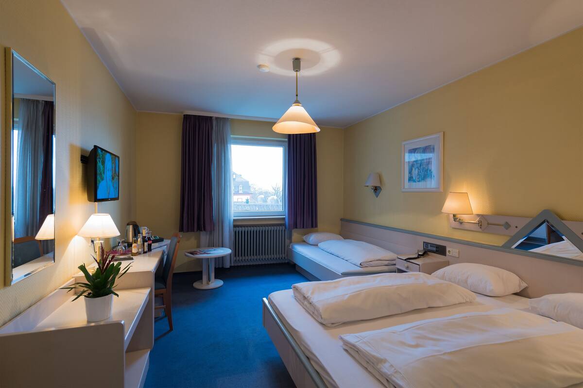 A hotel room with a bed, a desk, a chair, a small table, a potted plant, a window with curtains, a wall-mounted TV, and a painting.