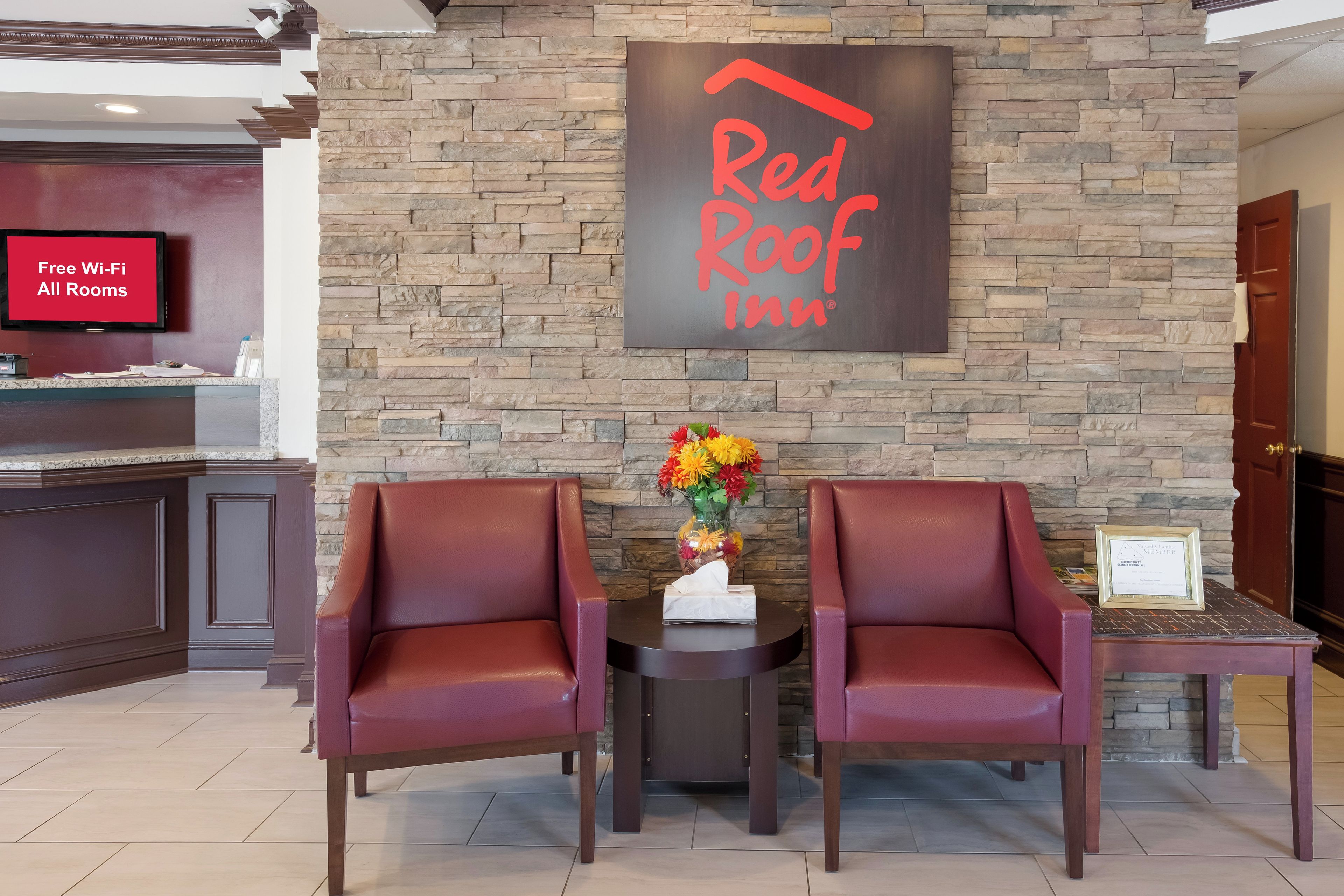 Zona de recepción de Red Roof Inn con un fondo de pared de piedra, dos sillones de cuero, un jarrón con flores y un cartel que indica Wi-Fi gratuito en todas las habitaciones.