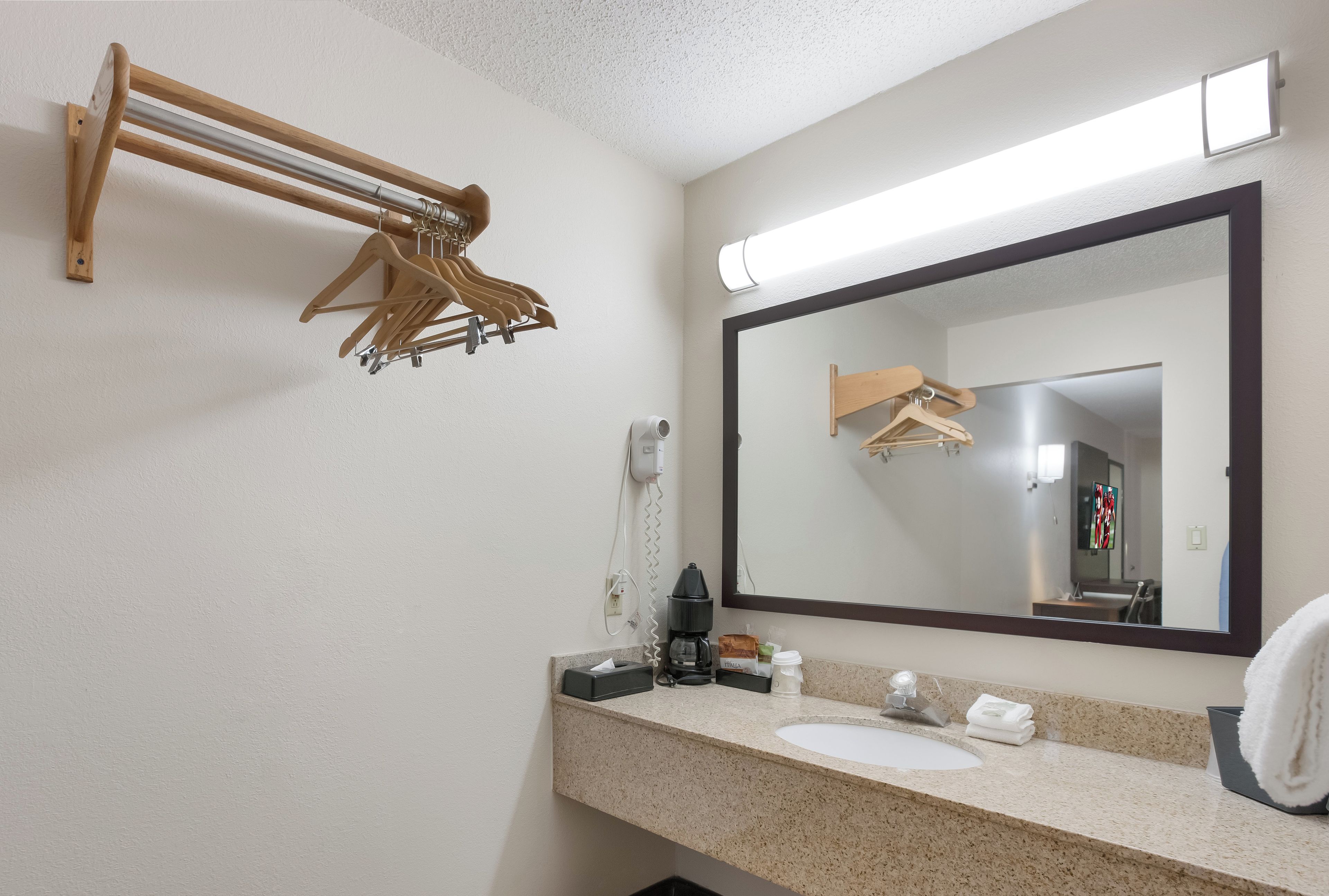 Une salle de bain d’hôtel équipée d’un porte-serviettes en bois, d’un grand miroir, d’un lavabo, d’une machine à café et d’un distributeur de serviettes.