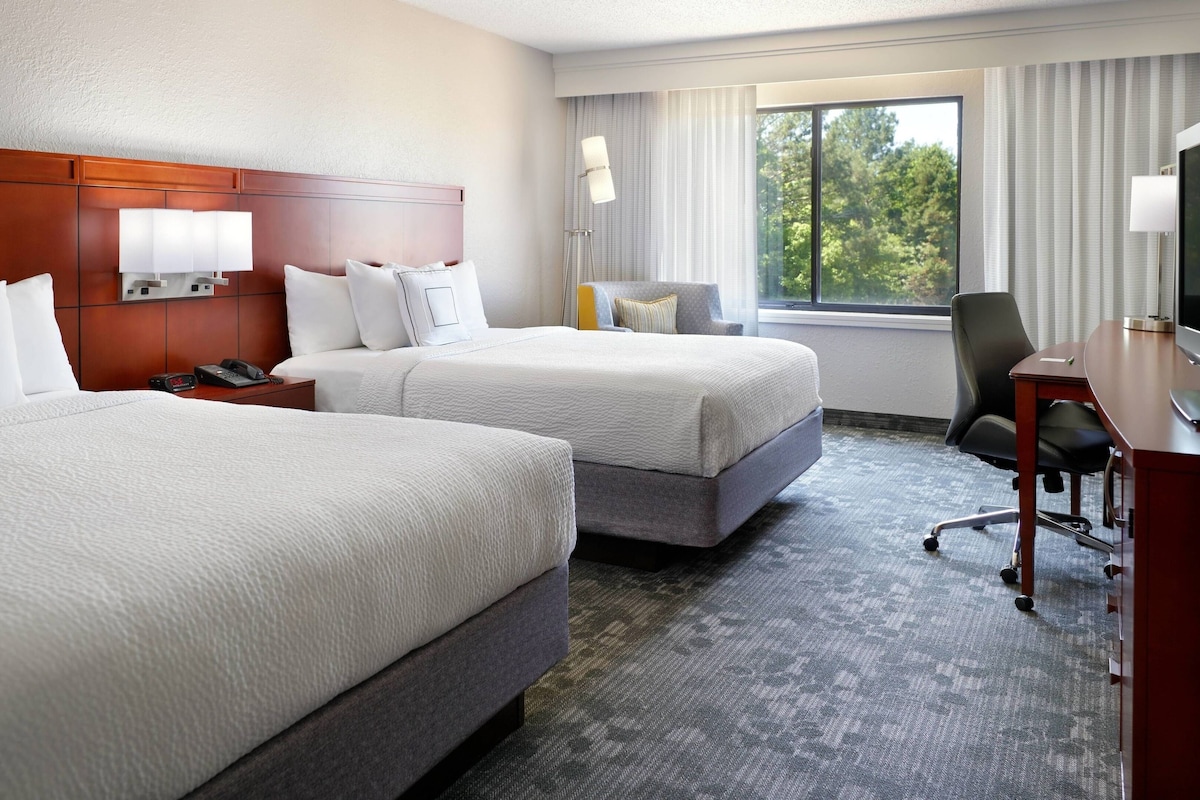 A hotel room with two beds, a desk with a TV, a chair, and a window with curtains.