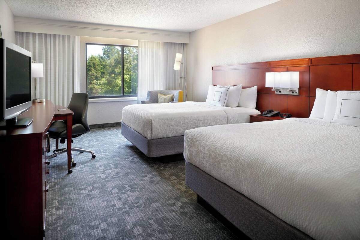 A hotel room with two beds, a desk with a TV, a chair, and a window with curtains.