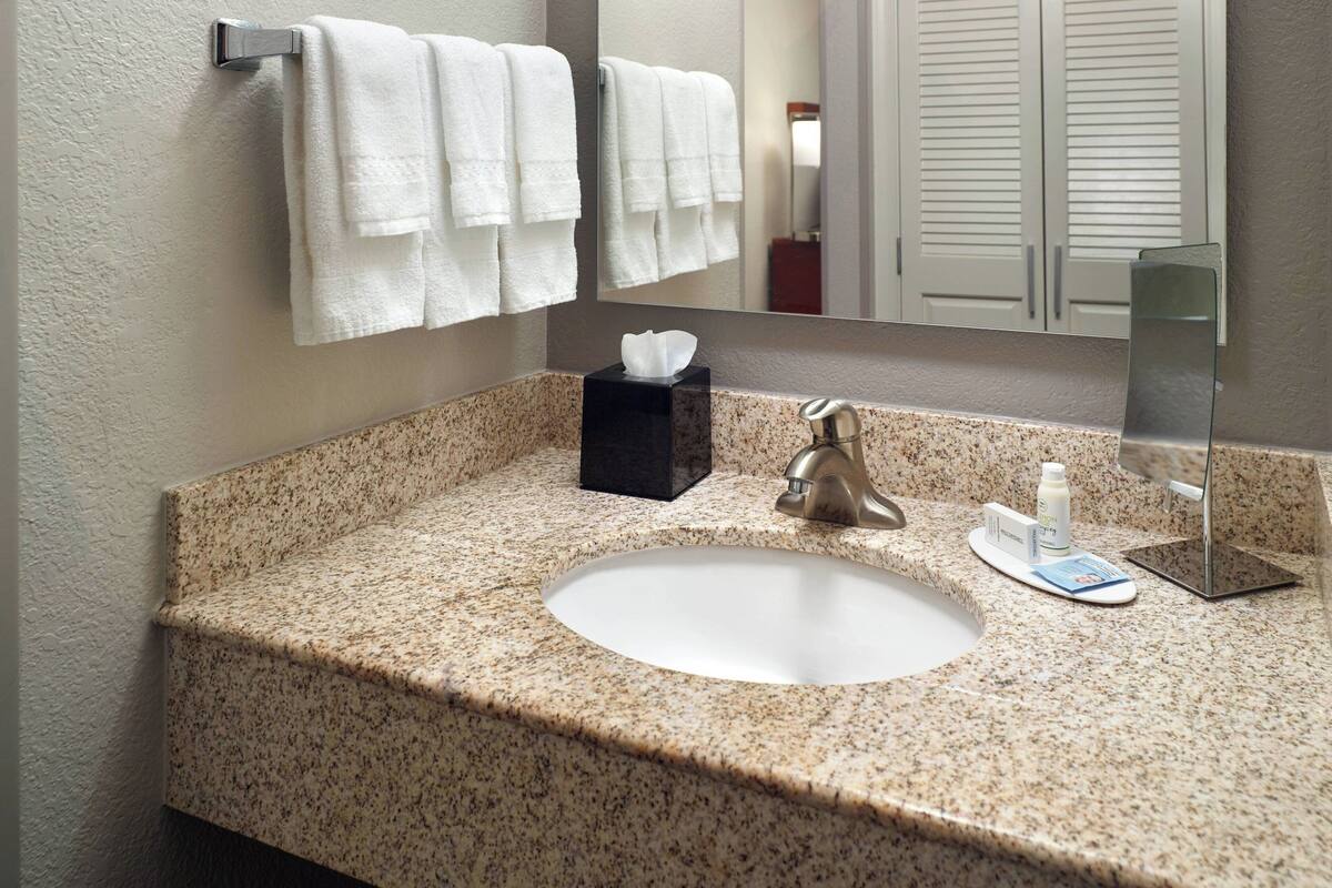 A bathroom with a granite countertop, a white sink, a towel rack with white towels, a tissue box, and a mirror.