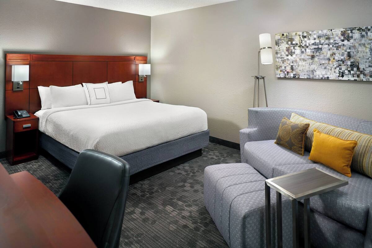 A hotel room with a large bed, a desk, a chair, a sofa with cushions, and a floor lamp.