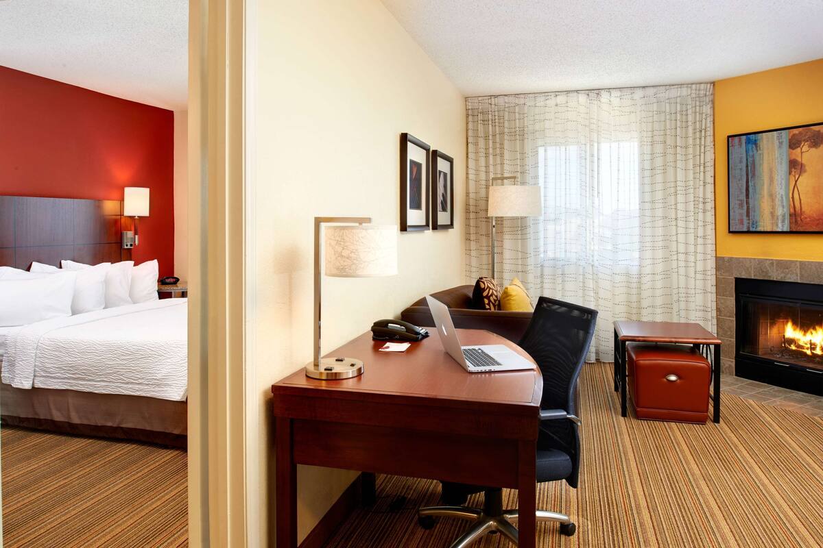A hotel room with a bed, desk, chair, lamp, fireplace, and a painting on the wall.