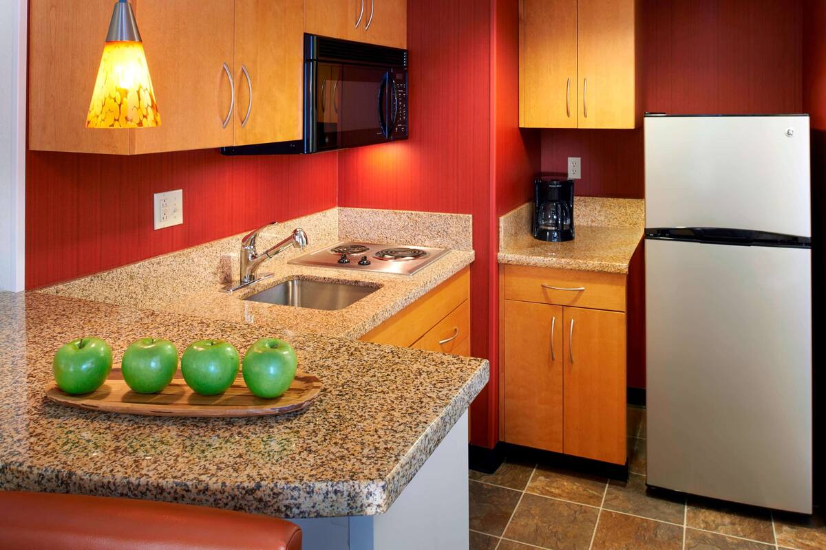 A kitchen with granite countertops, a stainless steel sink, a microwave, a stove, and a refrigerator. There are four green apples on a wooden tray on the counter.