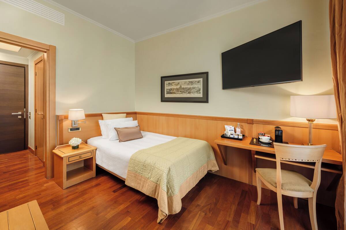 Una habitación de hotel con cama, mesita de noche con lámpara, escritorio con silla, televisor de pantalla plana y un cuadro en la pared.