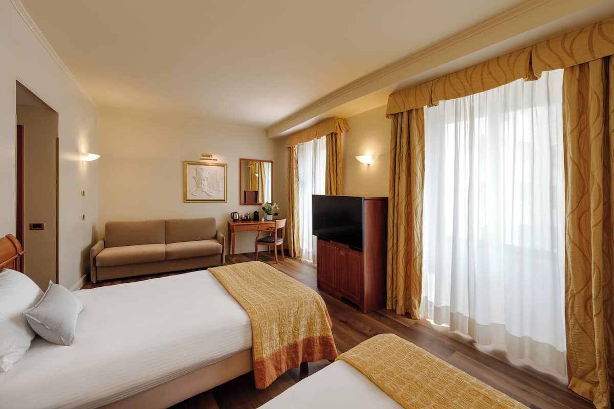Una habitación de hotel con dos camas, un sofá, un escritorio con televisor y ventanas amplias con cortinas.