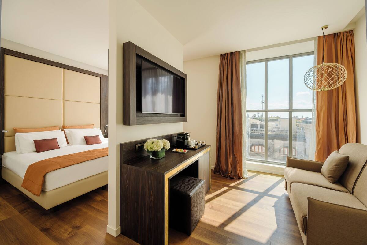 Habitación de hotel con cama, televisor montado en la pared, zona de estar con sofá y un ventanal con cortinas.