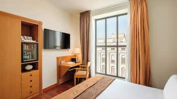 Una camera d'albergo con un letto, una scrivania, una televisione, un minibar e una finestra con tende.