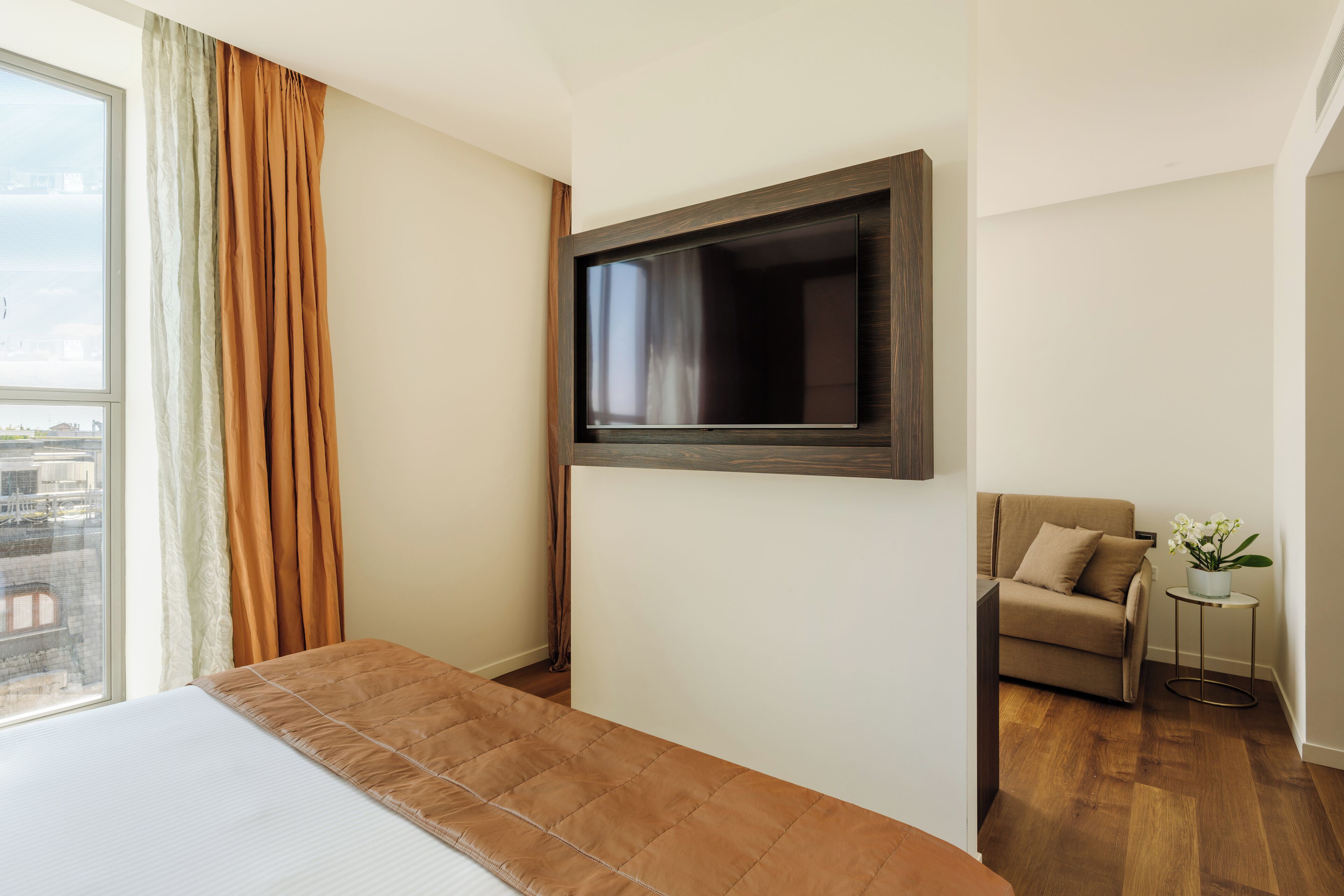 Ett hotellrum med en säng, en tv som är monterad på väggen, ett fönster med gardiner, ett litet bord med en växt och en soffa.