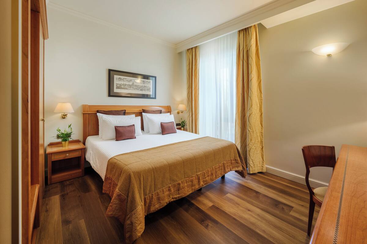 Habitación de hotel con una cama grande, cabecera de madera, dos mesitas de noche con lámparas, un cuadro enmarcado en la pared y una silla de madera.