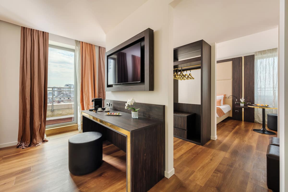 Una habitación de hotel moderna con un ventanal grande, un televisor de pantalla plana montado en la pared, un escritorio de madera oscura, un taburete redondo pequeño, una cama con cabecera blanca y una mesita con un jarrón de flores.
